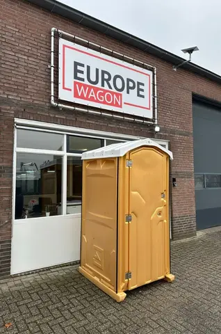 Een geel mobiel toilet staat voor een bakstenen gebouw met een bord waarop ‘Europe Wagon’ staat