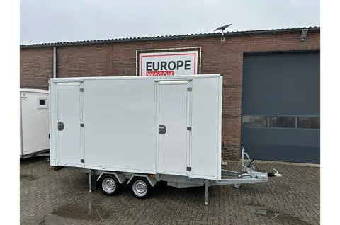 Een witte aanhangwagen met dubbele assen staat geparkeerd voor een bakstenen bedrijfsgebouw
