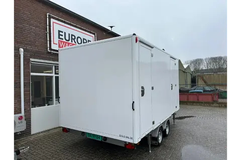 Een witte gesloten aanhanger met drie assen die geparkeerd staat voor een bakstenen bedrijfspand op een grijze dag