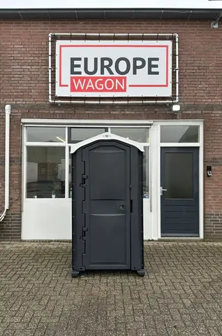 Een donkergrijze mobiele toiletcabine geplaatst voor de entree van een bakstenen pand