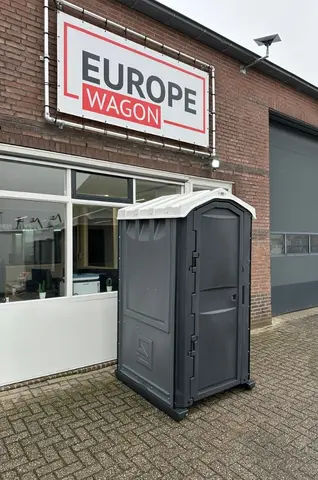 Een donkergrijze mobiele toiletcabine op een stoep naast een pand met een groot bord aan de muur