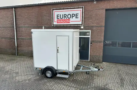 Een witte gesloten aanhanger staat geparkeerd voor een gebouw met het bord 'Europe Wagon'